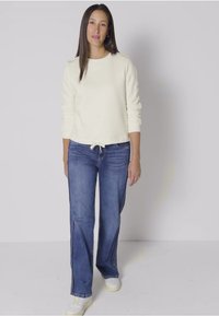 Krémový dlouhý rukáv s uzlem v pase, spárovaný s modrými zvonovými džínami a bílými teniskami. Jednoduchý, neformální design.