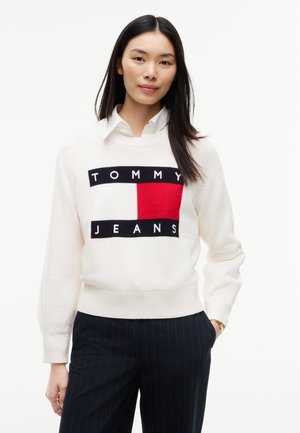 Junge Frau mit langen dunklen Haaren, die einen weißen Tommy Jeans Pullover und dunkle Nadelstreifenhose trägt und mit einer Hand in der Tasche steht.