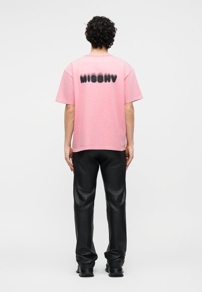 Άτομο με κατσαρά μαλλιά στέκεται με την πλάτη στραμμένη, φορώντας ροζ μπλουζάκι με μαύρο κείμενο "MISBHV" και μαύρο δερμάτινο παντελόνι και παπούτσια.