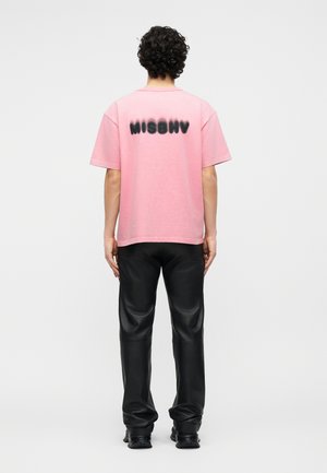 Osoba s kudrnatými vlasy stojí zády, má na sobě růžové tričko s černým nápisem "MISBHV" a černé kožené kalhoty a boty.