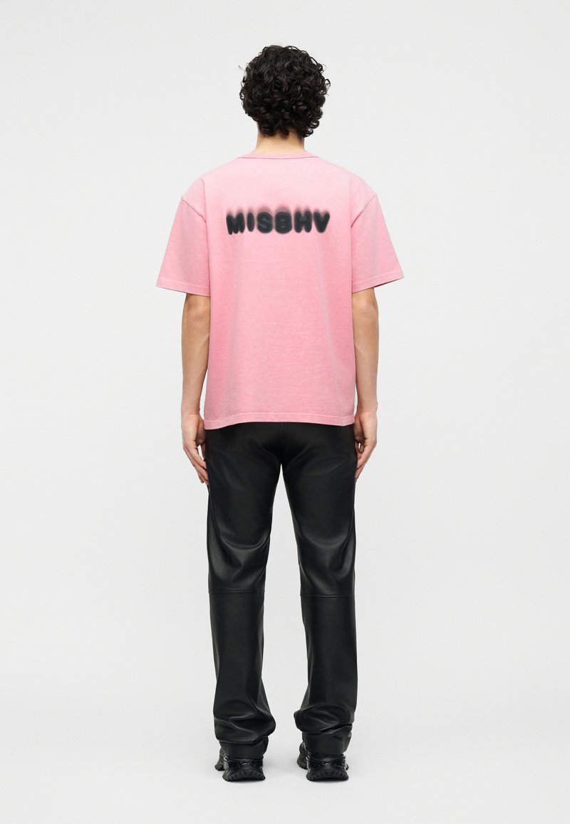 Persona con capelli ricci vista di schiena, indossa una maglietta rosa con la scritta nera "MISBHV" e pantaloni e scarpe di pelle nera.