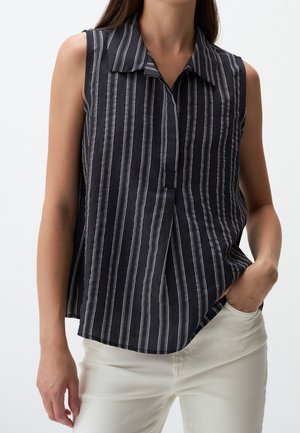 Chemise noire sans manches avec des rayures verticales blanches portée avec un pantalon blanc, une main dans la poche, sur un fond clair.