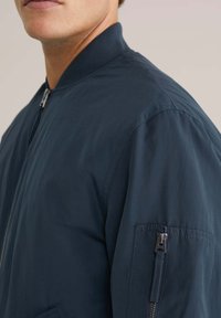 Giacca bomber blu navy realizzata in tessuto morbido e leggero. Presenta una tasca con cerniera sul braccio sinistro e colletto a costine.