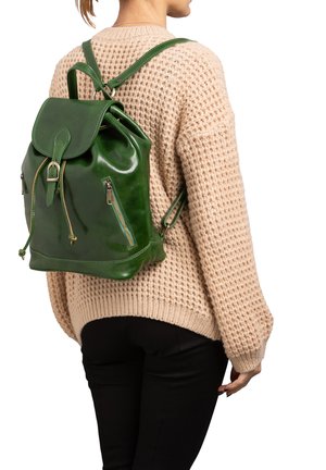 Vrouw draagt een beige gebreide trui en zwarte broek, met een groene leren rugzak met gespen en ritszakken op haar rug.