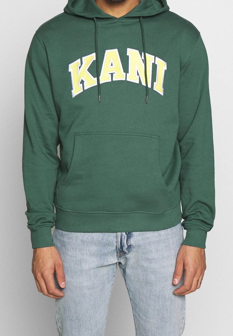 Grön bomullshuvtröja med ficka fram, prydd med "KANI" i gul och vit text på bröstet. Huva med justerbara snoddar.