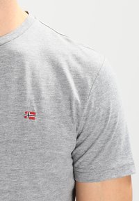 Šedé tričko s krátkými rukávy z měkké látky, s malou vyšívanou norskou vlajkou na hrudi. Viditelné detaily švů.