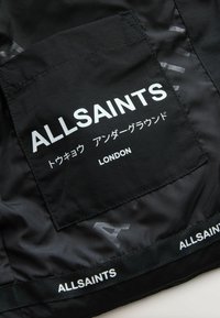 Svart tyg med en texturerad yta, med en stor ficka med "ALLSAINTS" i vitt, och ett dekorativt band vid fållen som också har texten "ALLSAINTS."