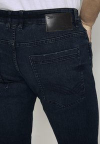 Tmavě modré džíny z denimu s zadní kapsou s výrazným vzorem šití a koženou nášivkou značky na pase.