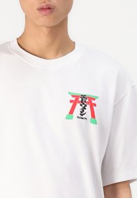Tricou din bumbac alb cu un grafic care ilustrează o poartă torii roșie și verde, cu caligrafie neagră, mâneci scurte și croială lejeră.