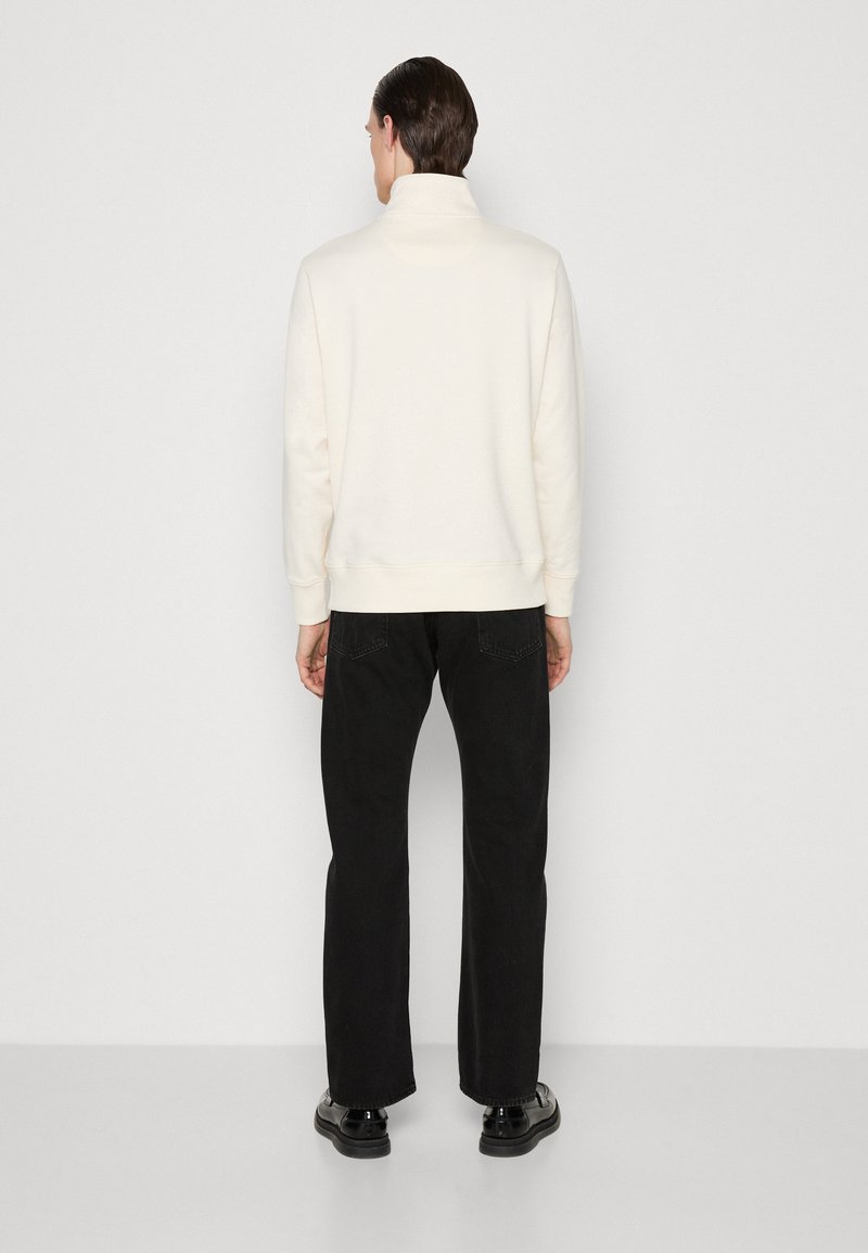 Uomo in piedi di fronte a un muro, indossa una felpa con collo alto color crema, jeans neri a gamba dritta e scarpe eleganti nere.