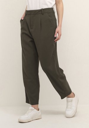 Femme portant un pantalon fuselé taille haute olive foncé avec plis, poches et baskets blanches, se tenant devant un fond uni.