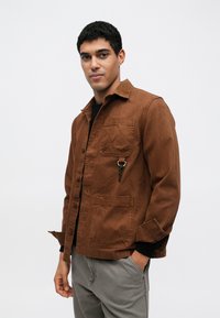 Pruun puuvillane jakk nööpidega esiküljel, rinnatasku ja võtmerõnga aksendiga. Disainitud lõdva lõike ja pikkade varrukatega.