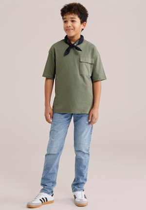 Junge steht, trägt ein olivgrünes T-Shirt mit Taschen, hellblaue Jeans, weiße Sneakers mit schwarzen Streifen und ein schwarzes Halstuch.