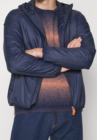 Giacca imbottita blu navy con cappuccio, chiusura lampo frontale, polsini elasticizzati e un tirante arancione, indossata sopra un maglione lavorato a maglia con sfumature.