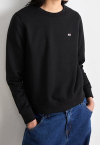 Sudadera negra con cuello redondo y puños acanalados, que presenta un pequeño logo colorido en el pecho y un corte relajado. Llevada con jeans azules.