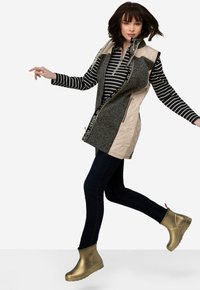 Lange grijze vest met beige accenten en ritsen, gedragen over een zwart-wit gestreepte top, gecombineerd met donkere jeans en metallic gouden enkellaarsjes.