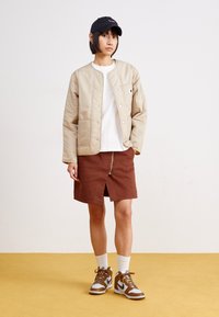 Beige tikandatud jakk nööpidega, valge T-särk, pruun lukuga seelik, valged lühikesed sokid ja pruunid spordijalatsid hallide aktsentidega.