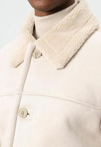 Beige jas met een zachte, gestructureerde faux shearling kraag, voorzien van een knoopsluiting en strakke lijnen. Zachte stof met een subtiele glans.