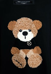 La tela negra presenta un oso de peluche texturizado en marrón y crema, con una camiseta negra y un detalle de logo circular en la camiseta.
