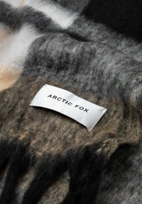 Couverture en laine avec un motif géométrique en noir, gris, beige et blanc, ornée d'une étiquette tissée "ARCTIC FOX". Surface douce et texturée.