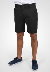 Schwarze Baumwollshorts mit geradem Schnitt, mit einem vorderen Knopfverschluss und seitlichen Taschen. Dazu werden navyblaue Wildledersneaker getragen.