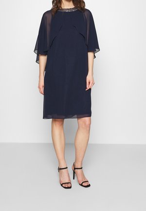 Kvinde iført en mørkeblå knælængde kjole med gennemskinnelige kappeærmer og udsmykket halsudskæring, kombineret med sorte højhælede sko med ankelrem.