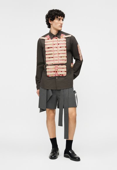 Mees kannab tumedat särki, mille peal on beežid rihmad punase lillemustriga riidel, sobitatud hallide seelikulaadsete lühikeste pükstega, millel on ripuvad riidetükid.