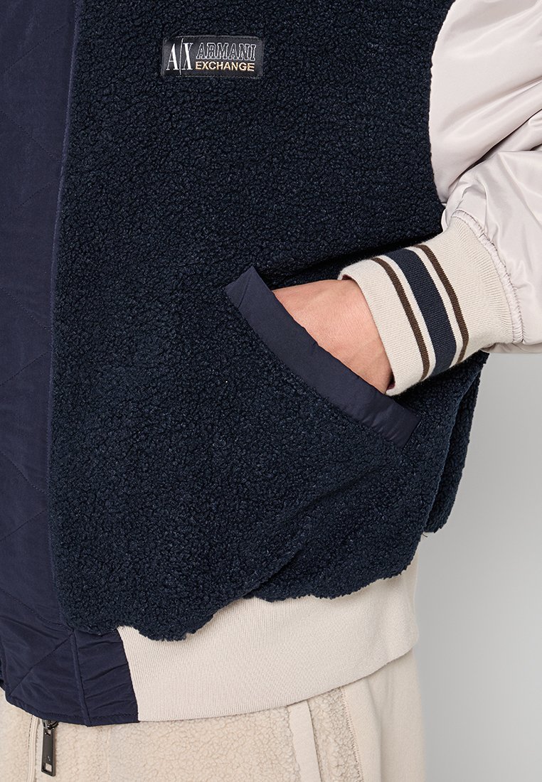 Giacca in fleece blu navy con tessuto trapuntato, polsini a righe a coste e tasca laterale. La patch con il logo sulla parte anteriore aggiunge un tocco di dettaglio.