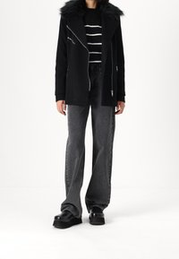 Chaqueta negra de mezcla de lana con cuello de piel sintética, cremalleras plateadas, combinada con unos vaqueros de pierna ancha grises y zapatos de plataforma negros. Suéter a rayas debajo.