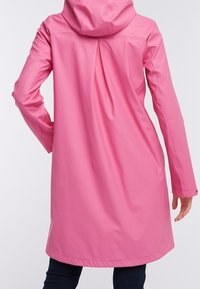 Donna che indossa un impermeabile rosa brillante, di lunghezza al ginocchio e con cappuccio, con maniche lunghe, in piedi con una mano sui fianchi, vista di spalle.