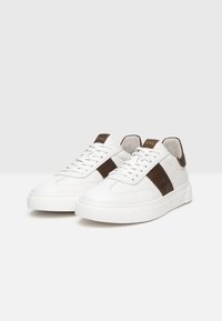 Sneakers bianchi in pelle con una striscia in suede marrone, lacci piatti e suola in gomma texture, con un logo sul tallone.