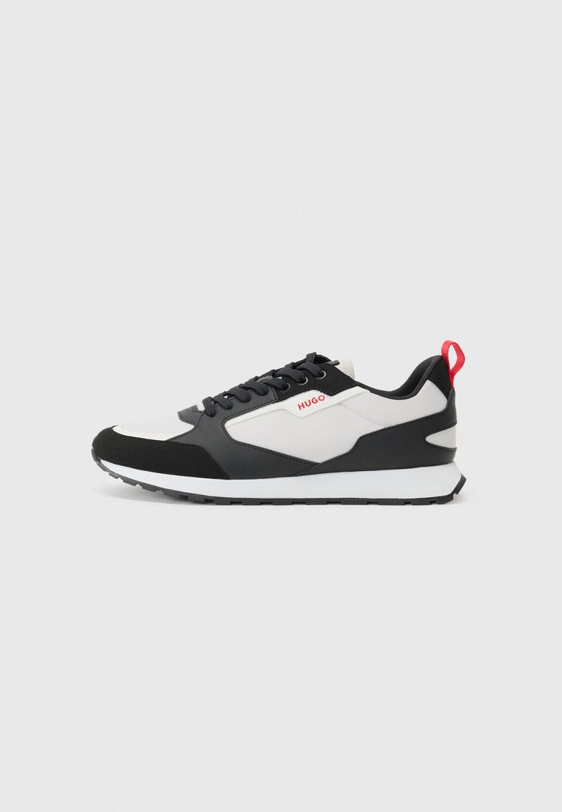 Black and white sneakers with a suede and mesh upper, featuring a red pull tab and textured sole. Lace-up design with logo detail.