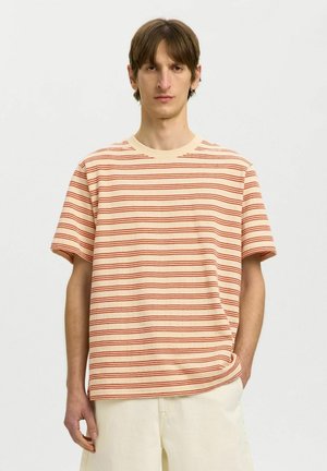Jeune homme aux cheveux bruns portant une chemise à manches courtes beige et orange rayée et un pantalon crème, debout devant un fond uni.