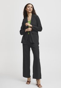 Schwarzes Blazer- und Hosen-Set aus leichtem Stoff, mit Reverskragen und vorderen Taschen, kombiniert mit einem gerippten grünen Oberteil und braunen Absatzschuhen.