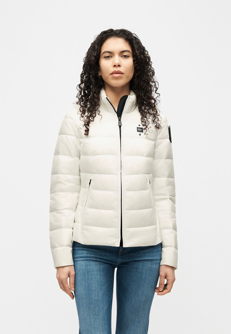 Γυναίκα με σγουρά μαλλιά που φοράει λευκό μπουφάν με φερμουάρ και μπλε τζιν, στέκεται μπροστά σε λευκό φόντο.