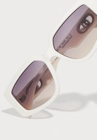 Hvide oversized solbriller med en kraftig ramme, gradient lila linser og indgraverede markeringer, der angiver materiale og dimensioner.
