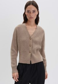 Femme avec de longs cheveux bruns portant un cardigan à boutons beige en côte et un pantalon noir, sur fond gris uni.
