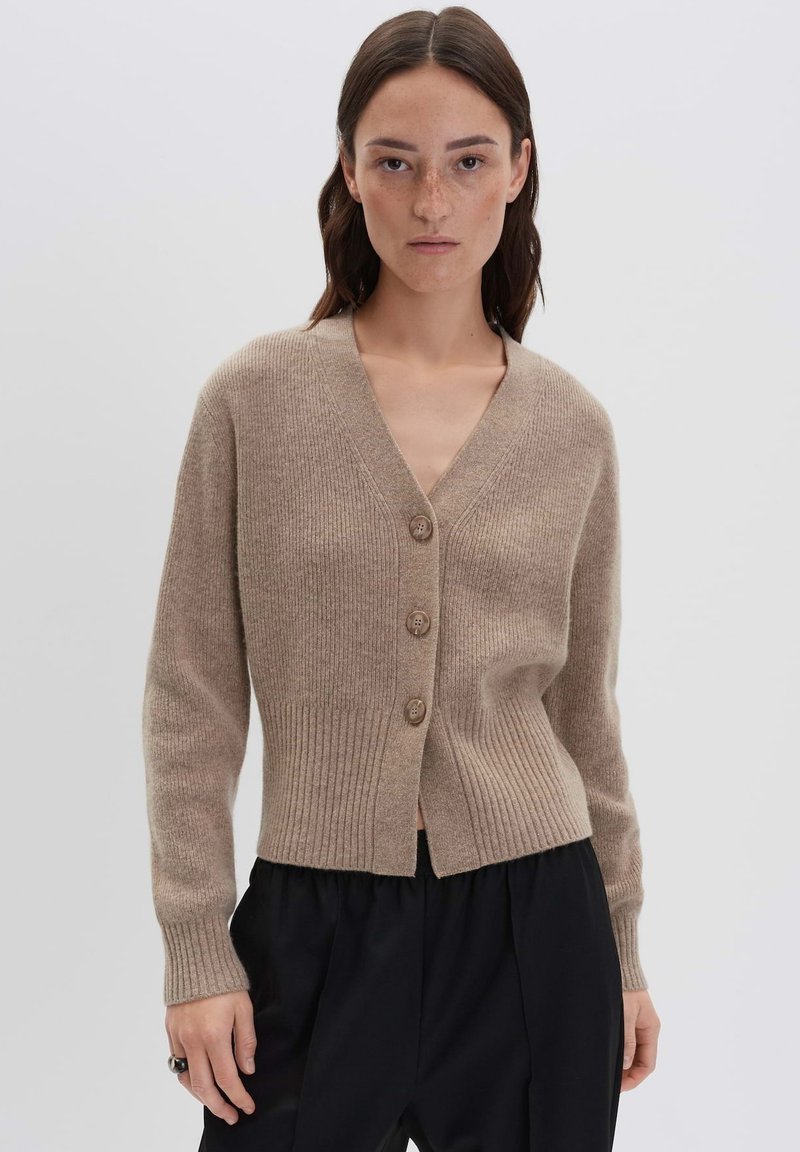 Femme avec de longs cheveux bruns portant un cardigan à boutons beige en côte et un pantalon noir, sur fond gris uni.