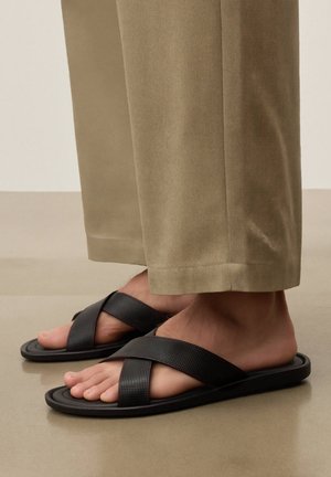 Lábak fekete, perforált, keresztpántos szandált viselnek, bézs színű, széles szárú nadrággal, sima padlón állva.