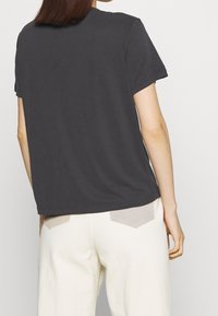 Mörkgrå bomullströja med korta ärmar och en avslappnad passform, tillsammans med krämfärgade byxor som har ljusa fickdetaljer.