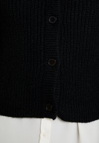 Schwarze Strickjacke mit geripptem Muster, ausgestattet mit fünf schwarzen Knöpfen an der Vorderseite, getragen über einem weißen Hemd mit Kragen.