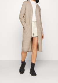 Bežový dlouhý kardigan vyrobený z měkkého pleteného materiálu, s otevřeným předním dílem, kapsami a žebrovanými manžetami. Nosený přes světlý top a lehké šortky s černými šněrovacími botami.