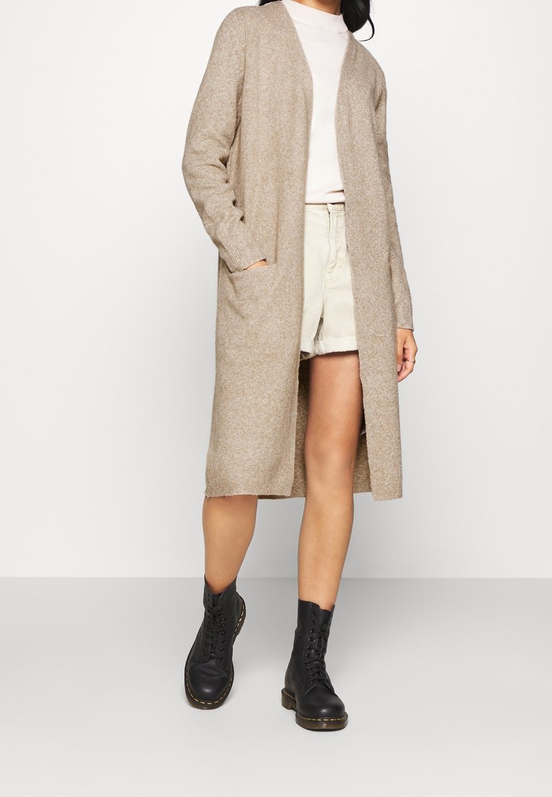 Bežový dlouhý kardigan vyrobený z měkkého pleteného materiálu, s otevřeným předním dílem, kapsami a žebrovanými manžetami. Nosený přes světlý top a lehké šortky s černými šněrovacími botami.