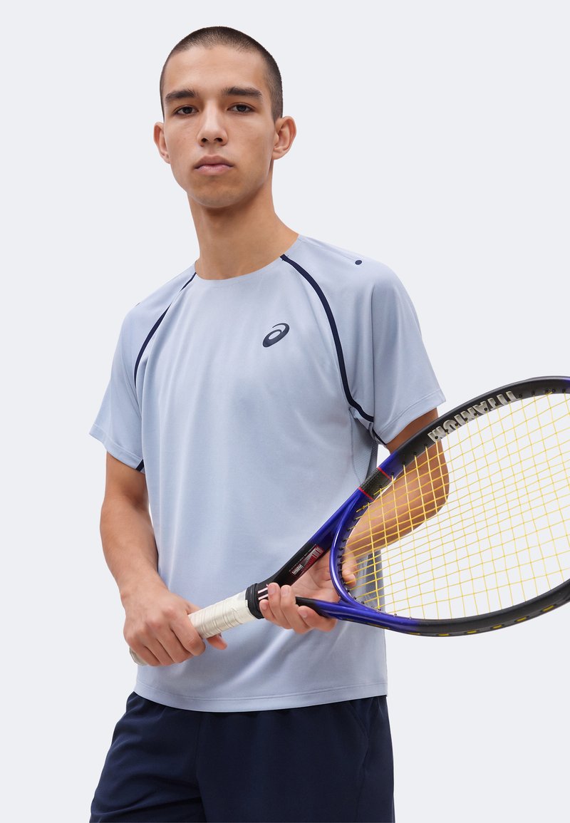 Ung mand, der holder en tennisracket, iført en lysegrå kortærmet sportstrøje og mørke shorts, stående foran en ensfarvet baggrund.
