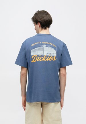 Fiatal férfi rövid barna hajjal, kék Dickies pólót visel sárga "Quality Workwear" felirattal és épület grafikával a hátán, bézs rövidnadrágban.