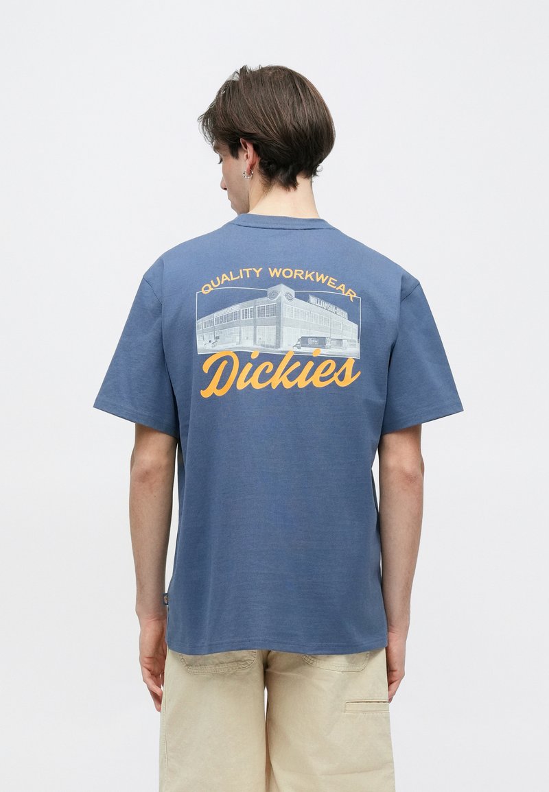 Junger Mann mit kurzem braunem Haar trägt ein blaues Dickies-T-Shirt mit gelbem "Quality Workwear"-Schriftzug und Gebäude-Grafik auf der Rückseite, beige Shorts.