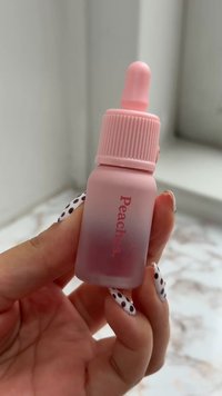 Una mano che tiene una piccola bottiglia contagocce rosa ghiacciata etichettata "Pesca", con unghie nere a pois visibili.