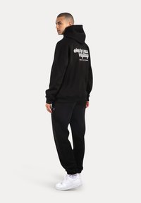 Felpa nera con cappuccio e vestibilità comoda, con testo bianco sul retro. Abbinata a pantaloni della tuta neri e sneaker bianche.