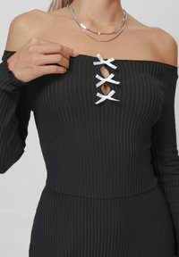 Svart ribbestrikket off-shoulder topp med nøkkelhull-detall og tre hvite bånd, lange ermer og en tettsittende silhuett.