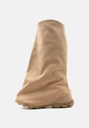 Botte haute beige en daim souple avec un bout arrondi et une semelle en caoutchouc, debout sur un fond blanc.